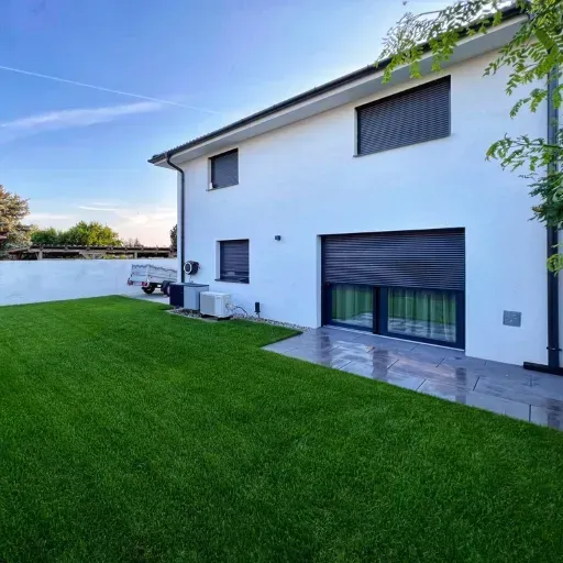 Casă modernă cu fațadă albă, rulouri exterioare din aluminiu și curte verde îngrijită.