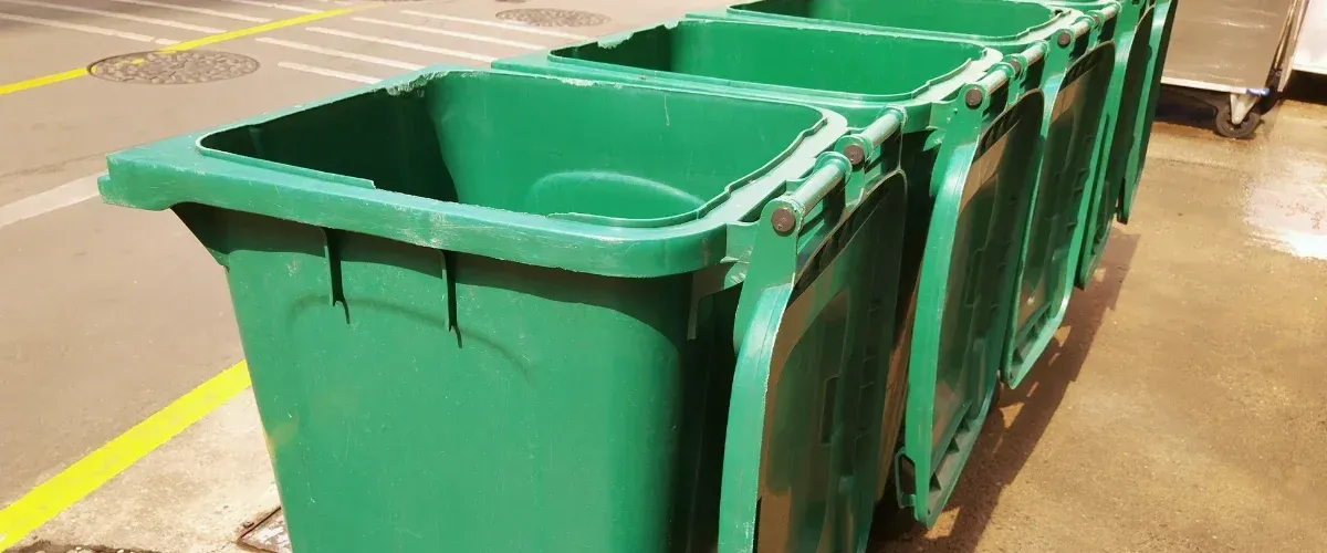 A row of four green recycling bins with open lids stand along a yellow-lined pavement on a sunny day.