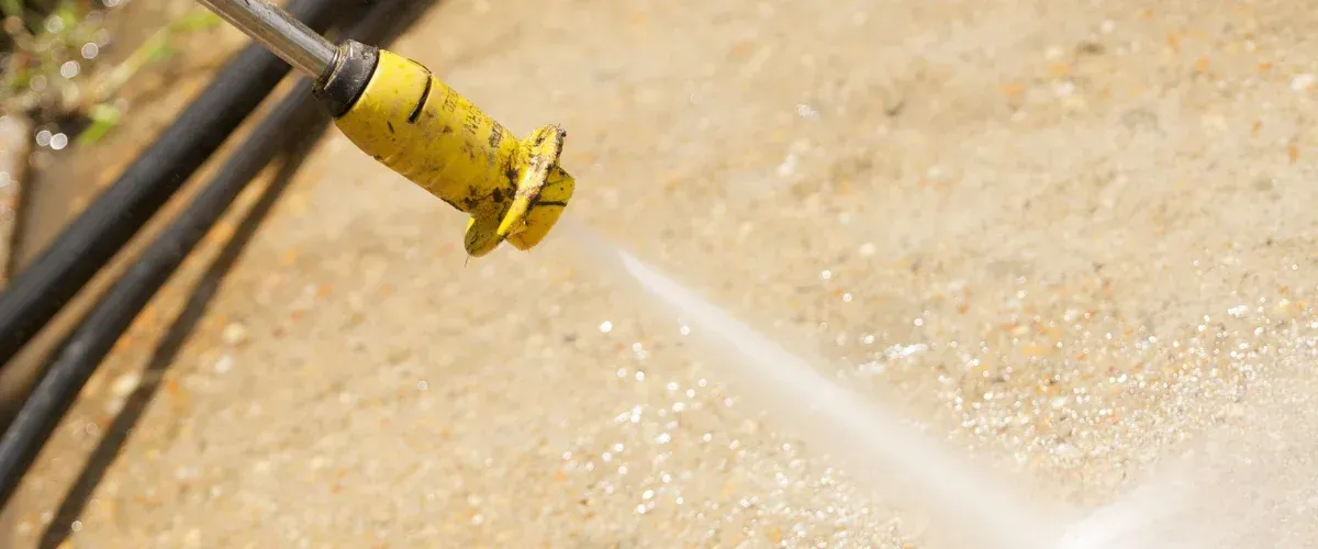 A close-up of a yellow pressure washer nozzle cleaning a speckled concrete surface with a stream of water.