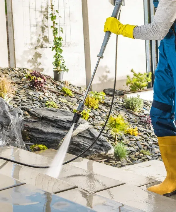 A person in yellow boots and gloves pressure washing patio steps outdoors, with a rocky garden in the background.