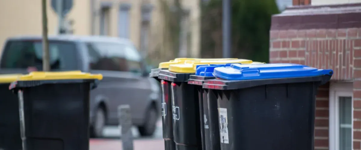 Multiple black recycling bins with yellow and blue lids lined up on an urban street next to a brick building.