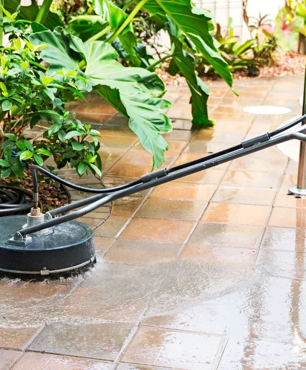 A close-up view of a circular surface cleaner attachment powered by a pressure washer cleaning a patio of square tiles.