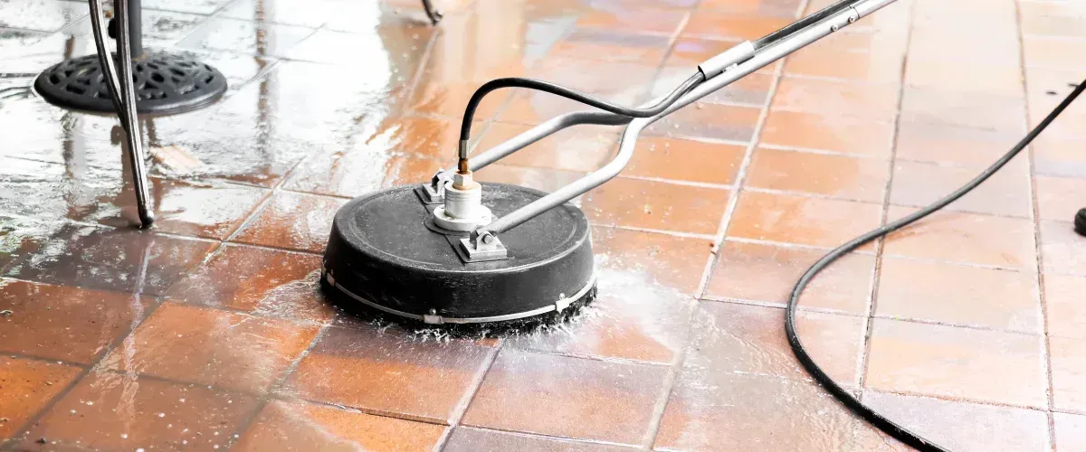A close-up of a yellow pressure washer nozzle cleaning a speckled concrete surface with a stream of water.