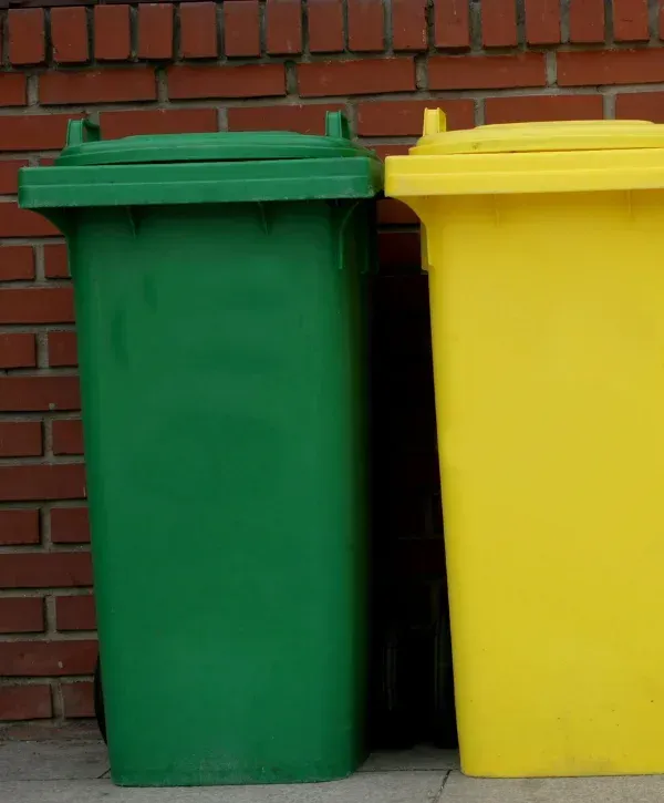 Two plastic recycling bins, one green and one yellow, standing side-by-side against a brick wall.
