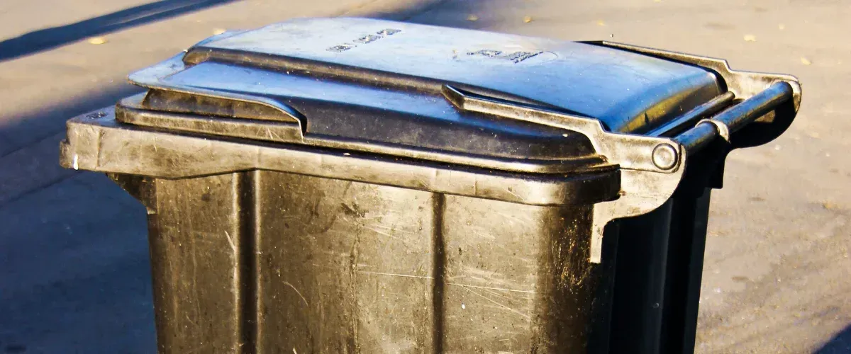 A close-up, high-angle shot of a dusty black recycling bin with its lid slightly ajar, sitting on asphalt.