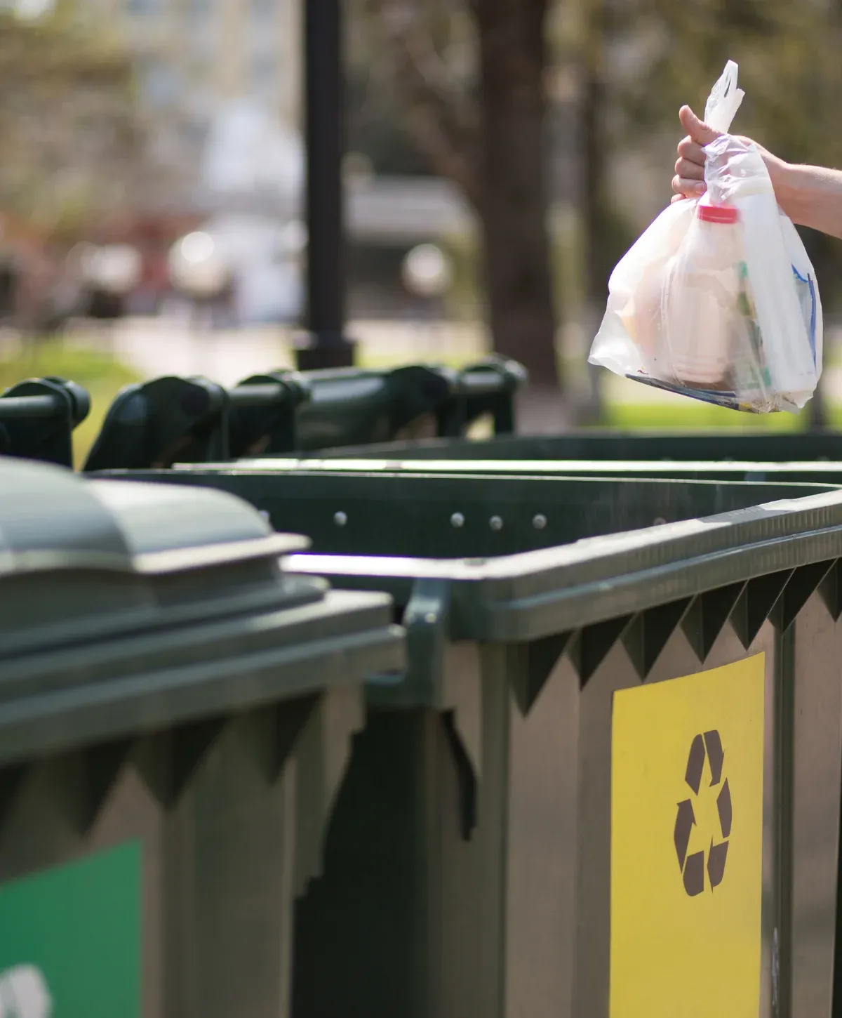 A hand holding a clear plastic bag of recyclables poised over a green outdoor recycling bin with a yellow symbol.