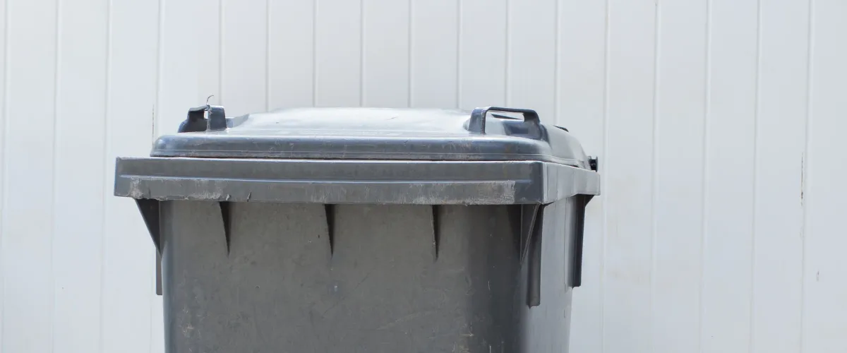 A weathered dark grey plastic trash can with a closed lid stands against a light grey slatted fence.
