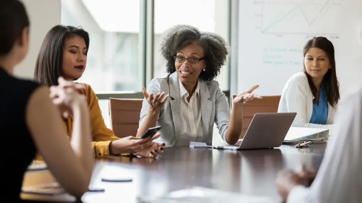    Five professionals sit around a conference table in discussion. One person at the center gestures while speaking, appearing to lead the conversation. Others listen attentively, with one holding a smartphone. A laptop, notebooks, and papers are on the table. In the background, a whiteboard shows a line graph and notes about growth comparison, a 2023 forecast, and sales growth.