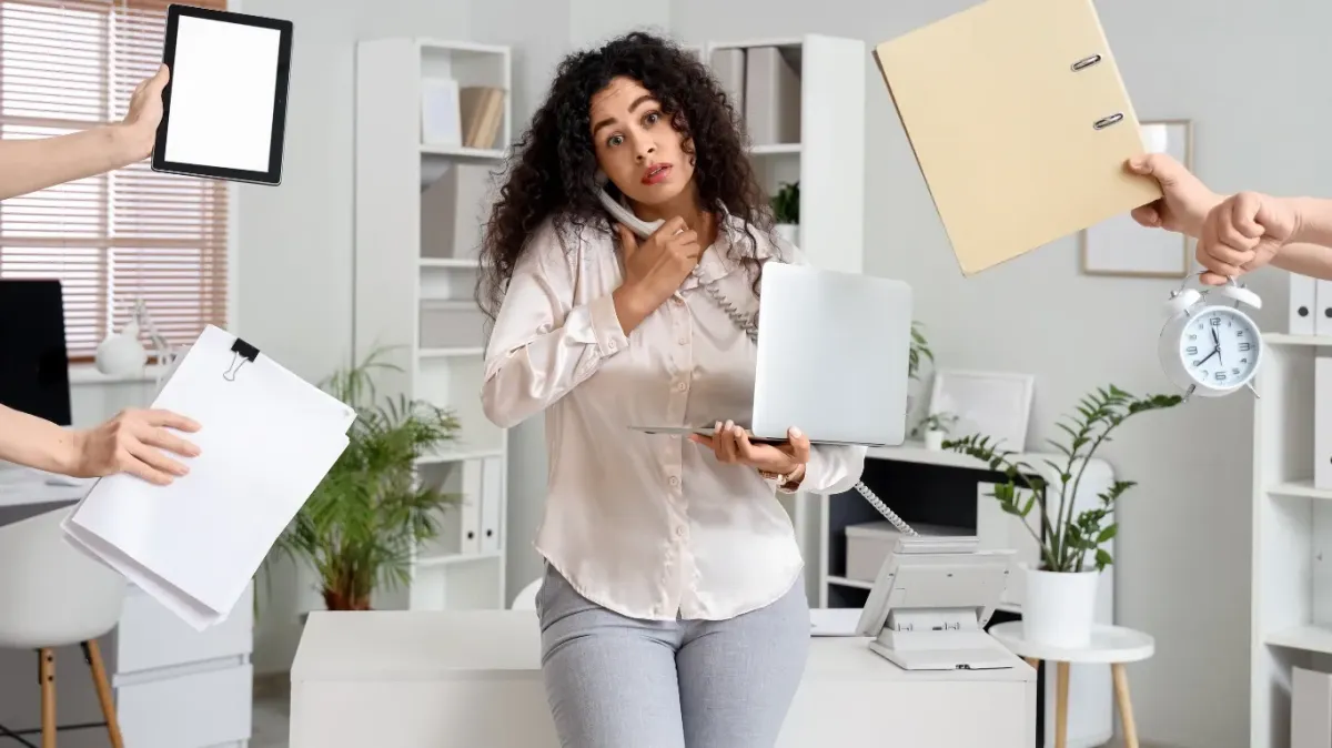 Overwhelmed professional woman multitasking in an office, illustrating causes of stress and practical ways to manage it, with competing demands like phone calls, deadlines, paperwork, and constant interruptions.
