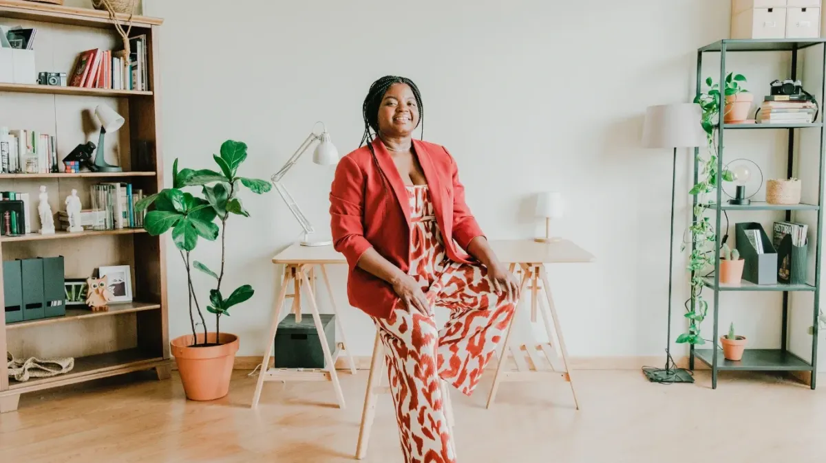 A smiling Black woman in a red blazer and patterned jumpsuit sitting in a bright, modern home office with plants and bookshelves, embodying Energy-smart Leadership Mantras for Managing Capacity.