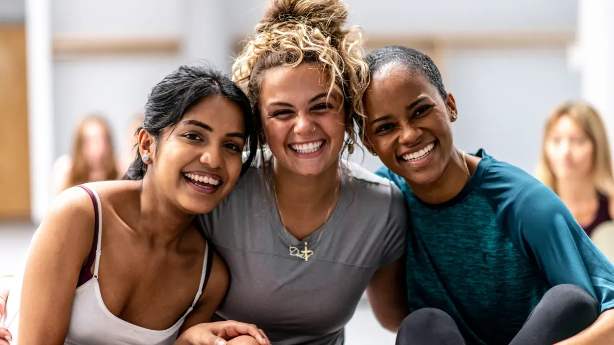 Three diverse women smiling together in a supportive, uplifting moment, reflecting women’s health prioritization and why your wellbeing comes first through connection, confidence, and community.