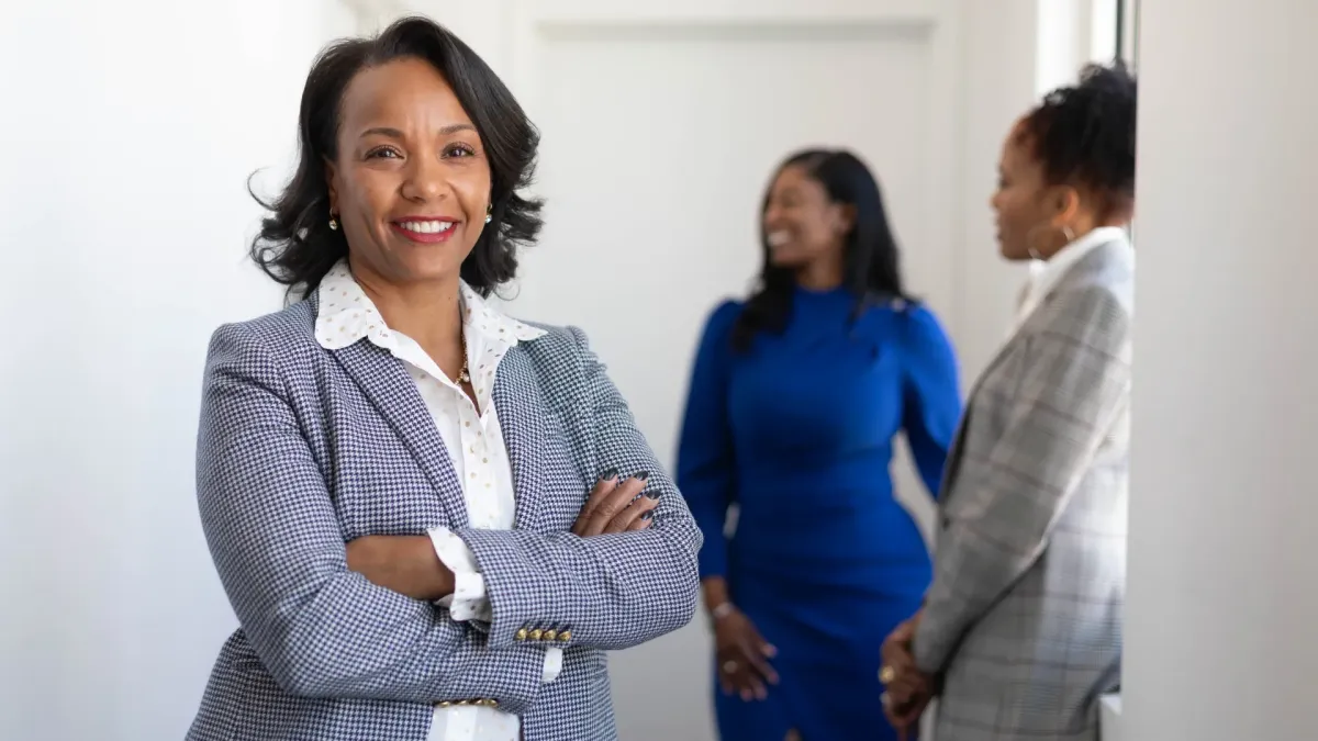 A confident businesswoman standing with arms crossed and smiling in a professional setting, with colleagues conversing in the background, illustrating decision-ready leadership mantras for confident choices, clarity, and self-assured leadership presence.