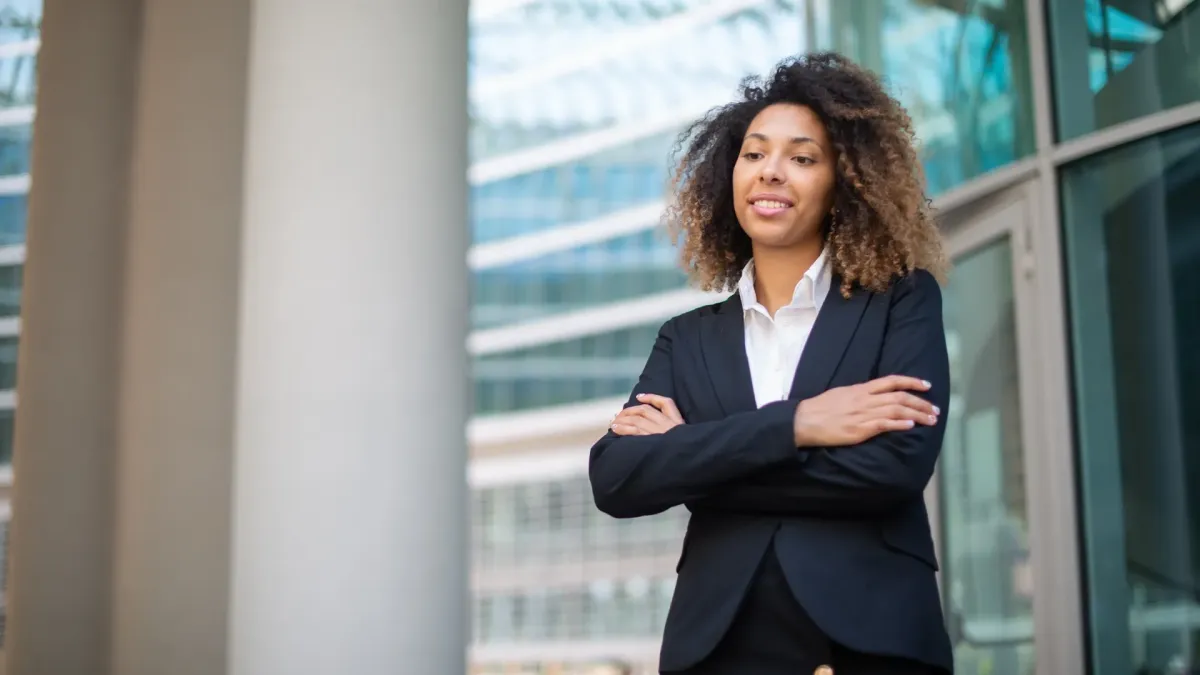 A confident professional woman standing with arms crossed outside a modern office building, representing people-first leadership mantras for team well-being, supportive leadership, and fostering a positive, balanced work environment.