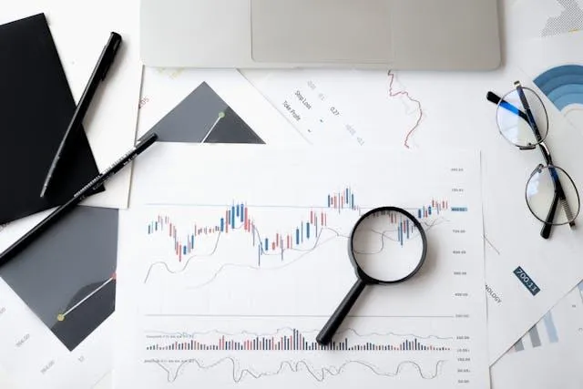 Graphs and charts on paper with a magnifying glass and a laptop, illustrating data analysis and performance monitoring for business optimization.