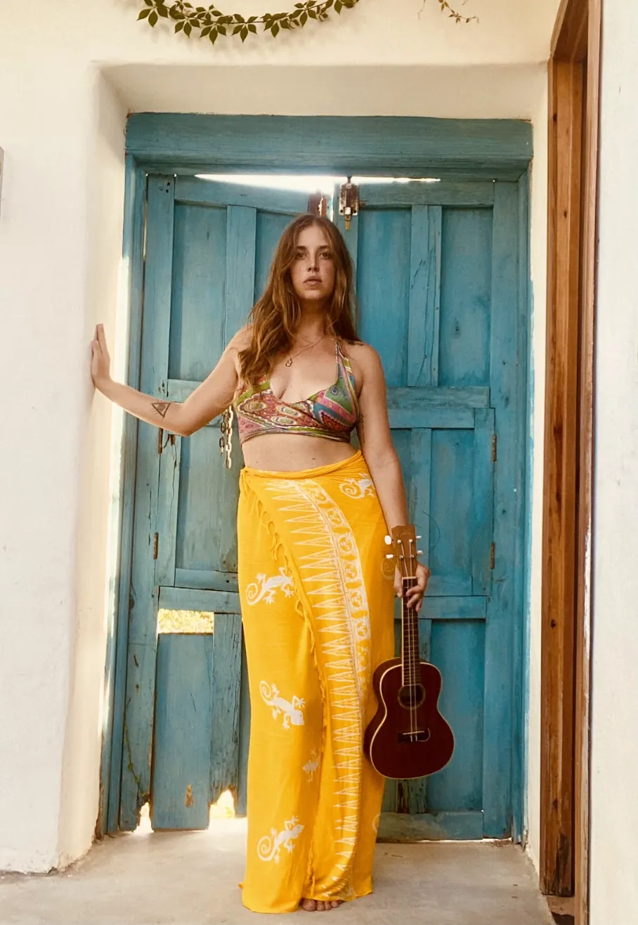 A smiling woman in her 30s with curly hair, wearing a soft pink blouse, seated at a piano with sheet music, surrounded by gentle natural light and a cozy, creative studio vibe.