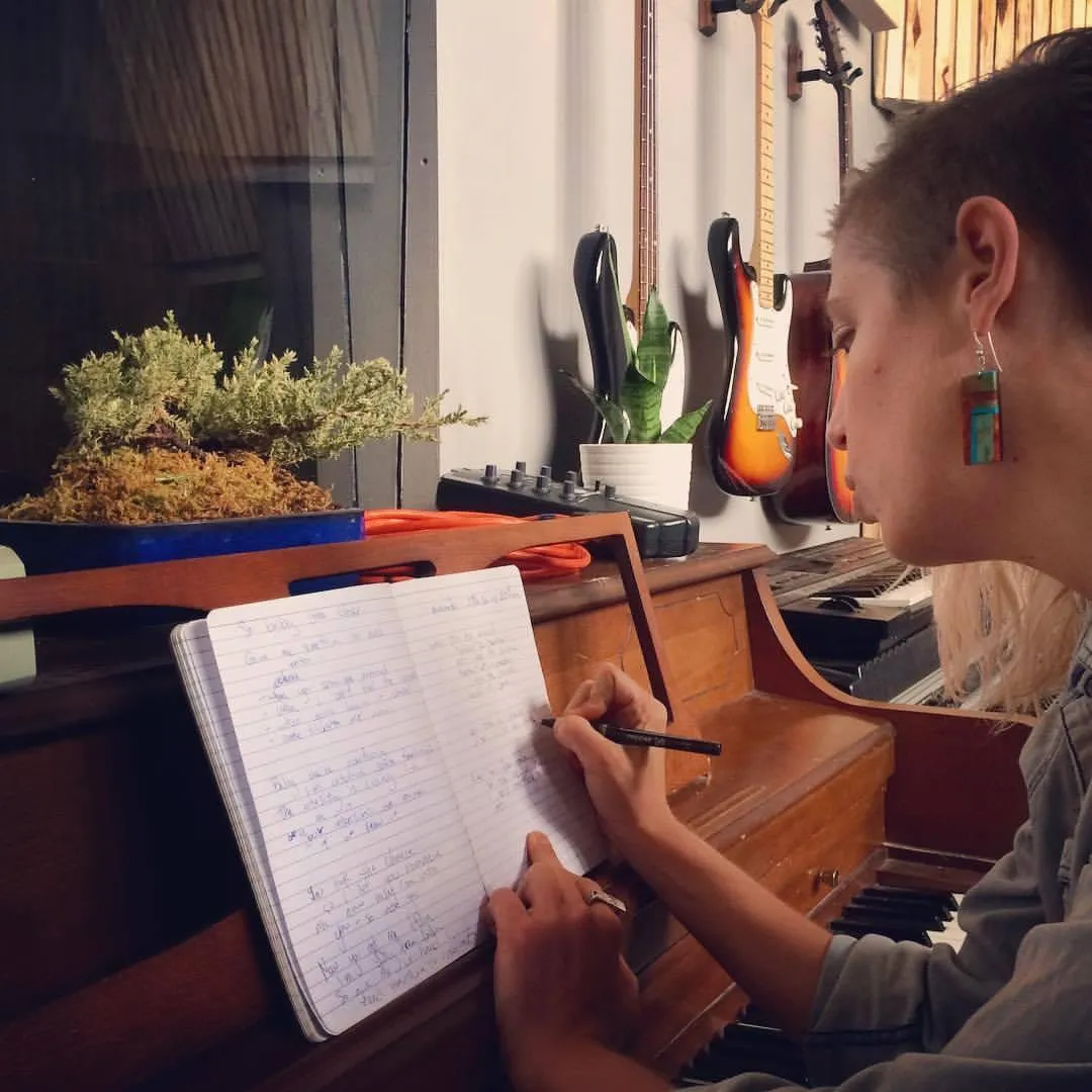 A young woman with headphones, smiling as she writes lyrics in a sunlit studio, surrounded by musical notes and soft pink decor, radiating creative energy and joy.