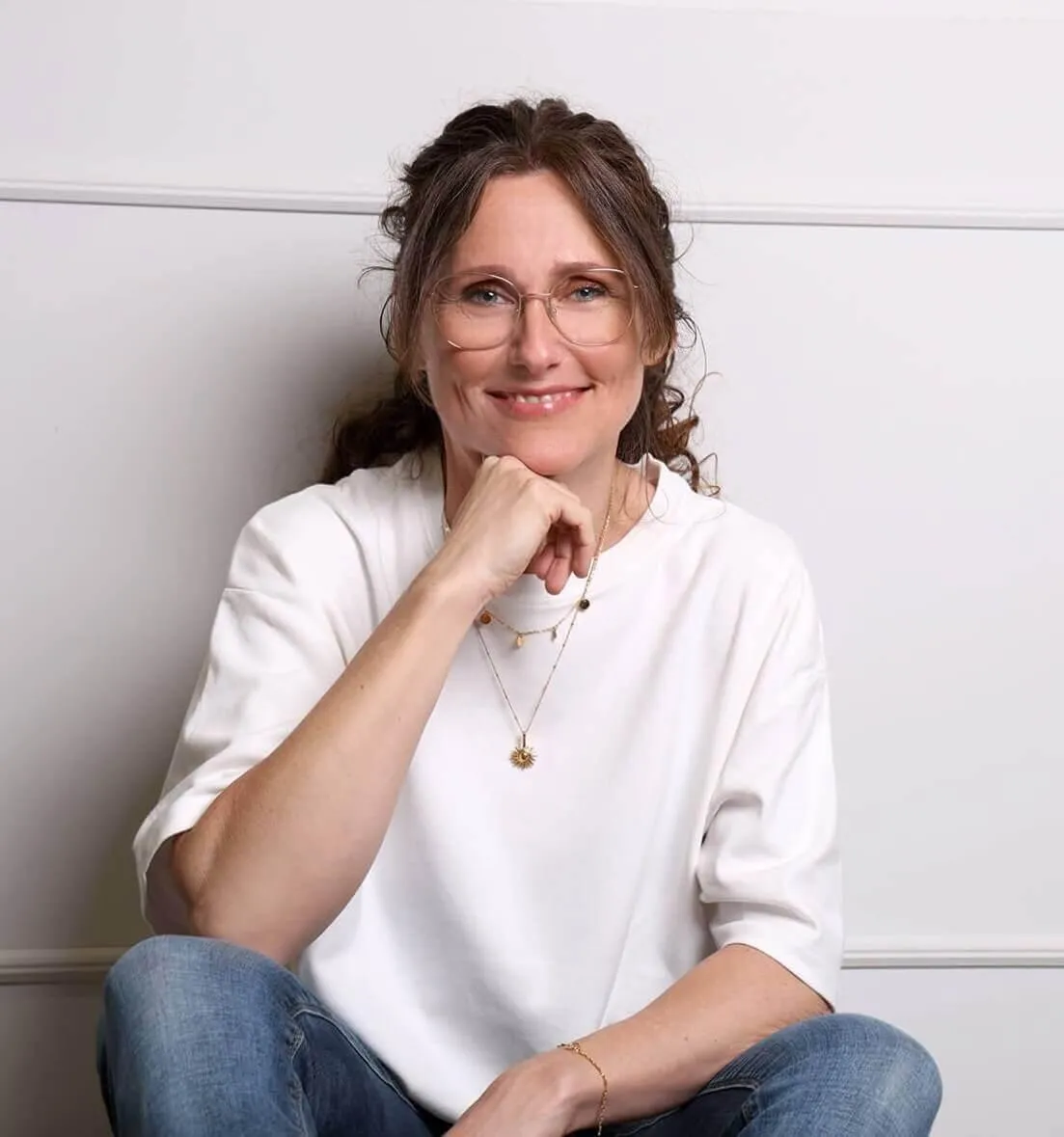 Foto Anja Glashörster, sitzend, weißes T-Shirt, blaue Jeans, den Kopf auf die Hand gestützt
