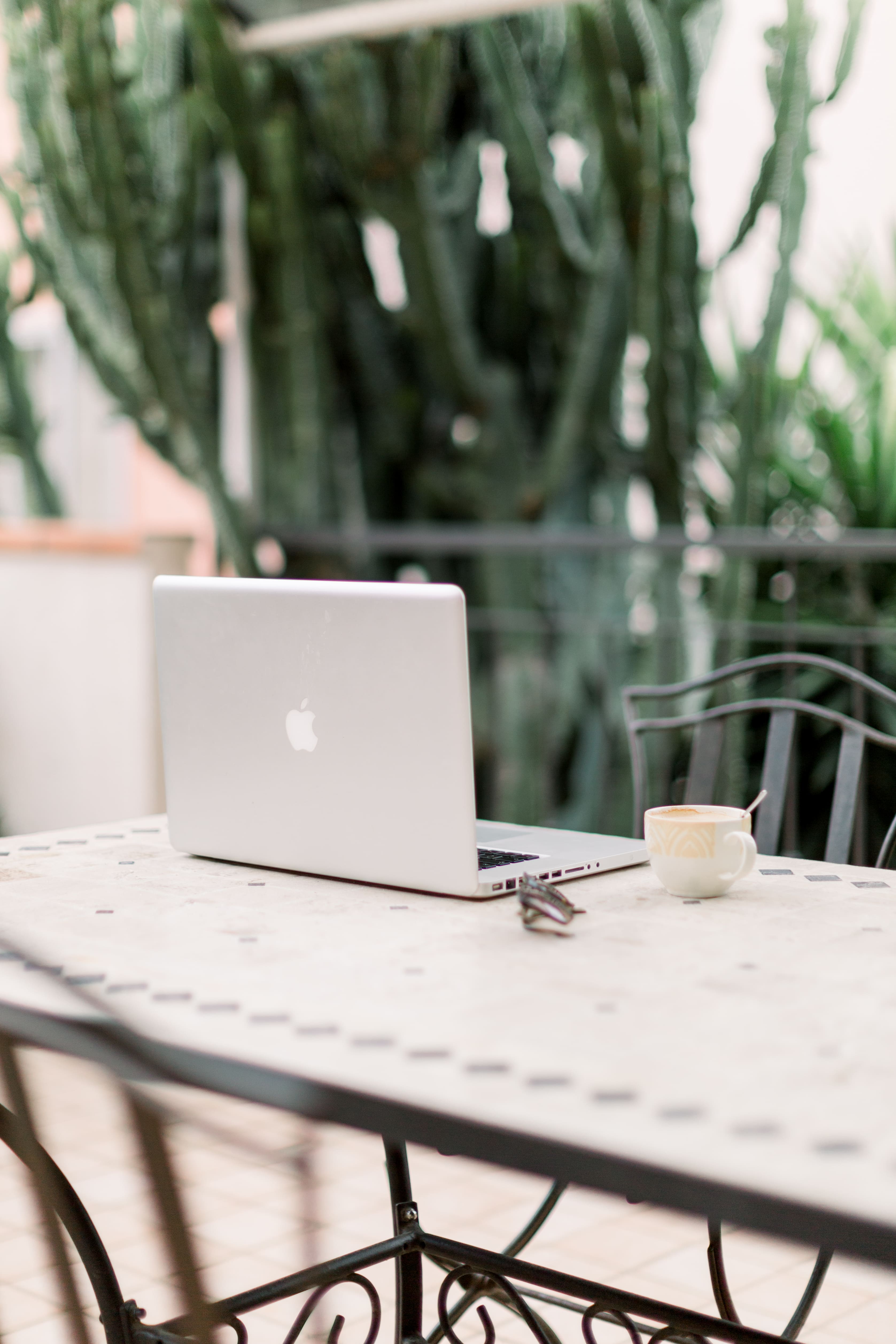 Laptop auf verschnörkeltem Tisch, Kaffeetasse + Sonnenbrille. Pflanzen im Hintergrund