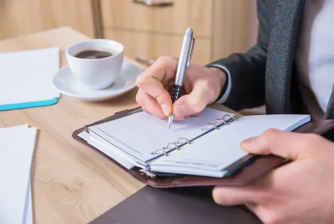 Man writing in a ring boun notepad at a desk with a cup of coffee