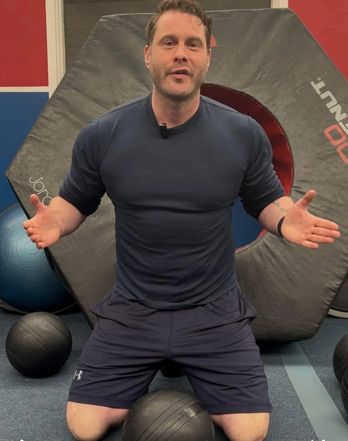 A friendly, multi-ethnic team of three fitness professionals stands together in a bright studio, smiling and holding clipboards and water bottles, ready to assist clients. The setting is modern and welcoming.