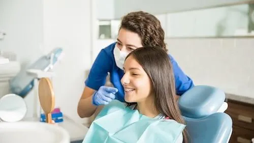 An orthodontist in a mask and gloves points at a young woman’s teeth as she smiles in a dental chair