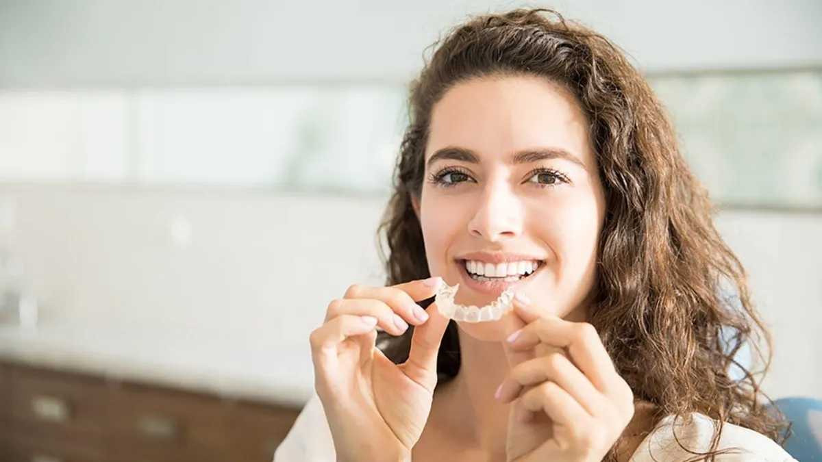 A young lady with long curly hair smiles, holding a clear dental aligner close to their mouth.