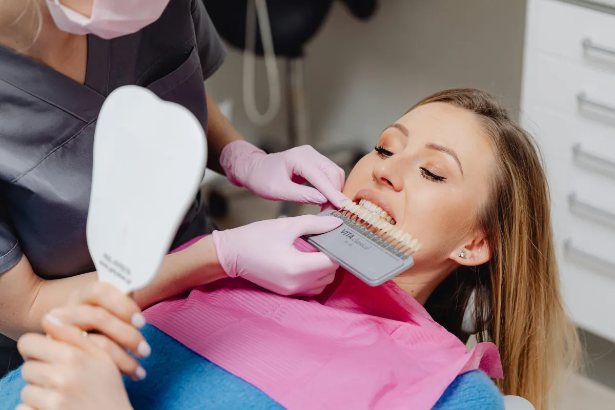 A woman in a dental chair has her tooth color matched with a shade guide by a dental professional.