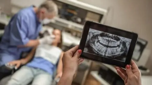 A orthodontist examines a patient while holding an X-ray on a tablet.