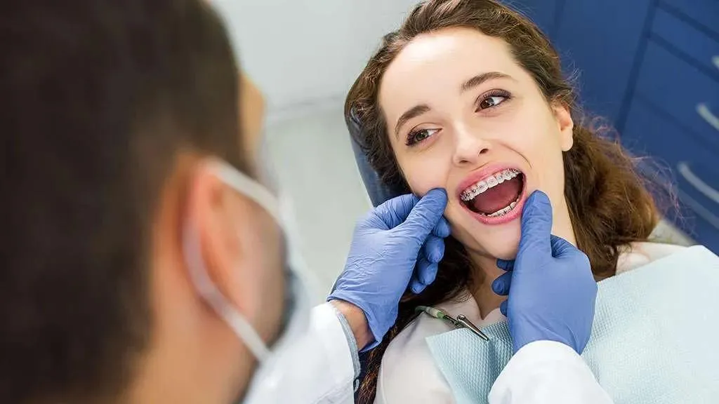 An orthodontist in gloves points at a young woman’s braces teeth as she smiles at him