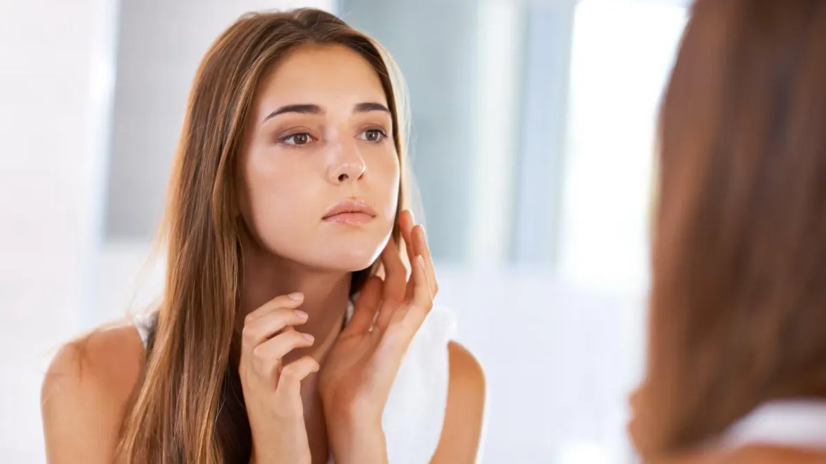 A woman with long brown hair touches her face, gazing at her reflection in a dentist office.