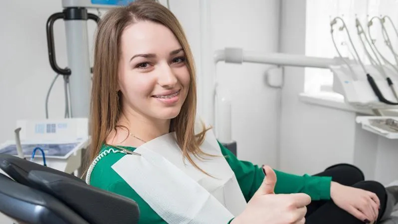 A woman with long hair smiles and gives a thumbs-up in a dental chair, with orthodontic equipment in the background.