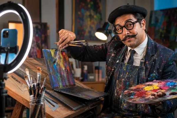 Agente de seguros disfrazado de pintor excéntrico —boina, bigote tipo Dalí y traje manchado de pintura— pintando la pantalla de una laptop frente a un aro de luz con un celular: crear contenido para redes sin ser diseñador.