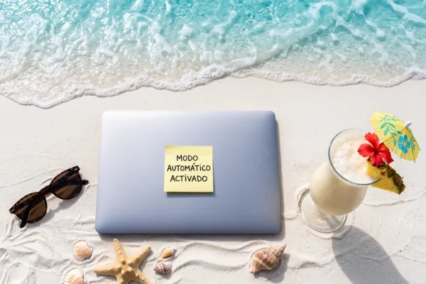 Laptop cerrada sobre la arena de una playa con un post-it que dice "Modo automático activado", junto a gafas de sol, una piña colada y conchas marinas, con la orilla del mar al fondo