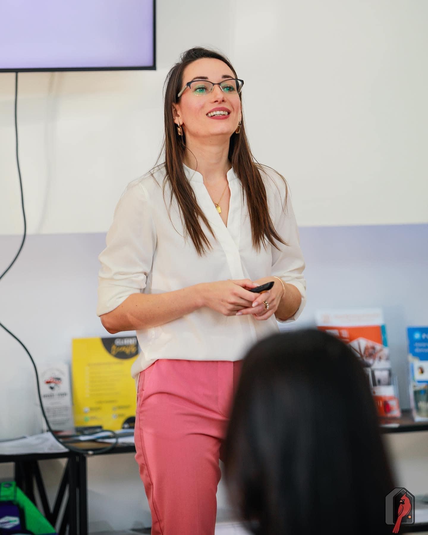 A woman wearing glasses, a white blouse, and pink pants stands at the front of a room giving a presentation. She is smiling and holding a small remote in her hands. A screen is mounted behind her, and informational materials are displayed on a table nearby. An audience member’s head is visible in the foreground, slightly out of focus.