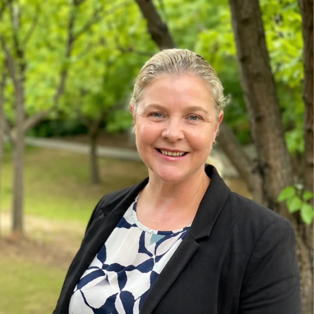 Professional woman smiling in a park setting, wearing a blazer and patterned blouse, representing career coaching for APS professionals in Canberra.