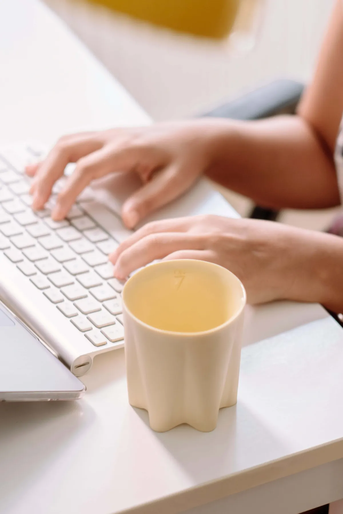 Hands typing at a white keyboard with a empty mug