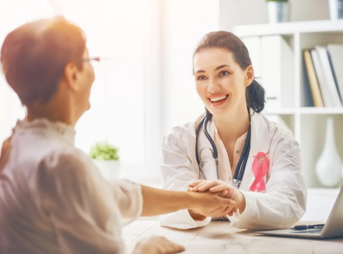 female doctor wearing a pink ribbon holding the hand of her aging female patient and smiling