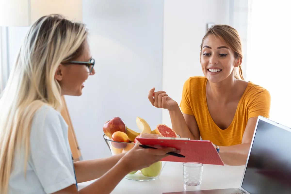 two women sitting at a white counter with a laptop, a bowl of fruit and a clipboard