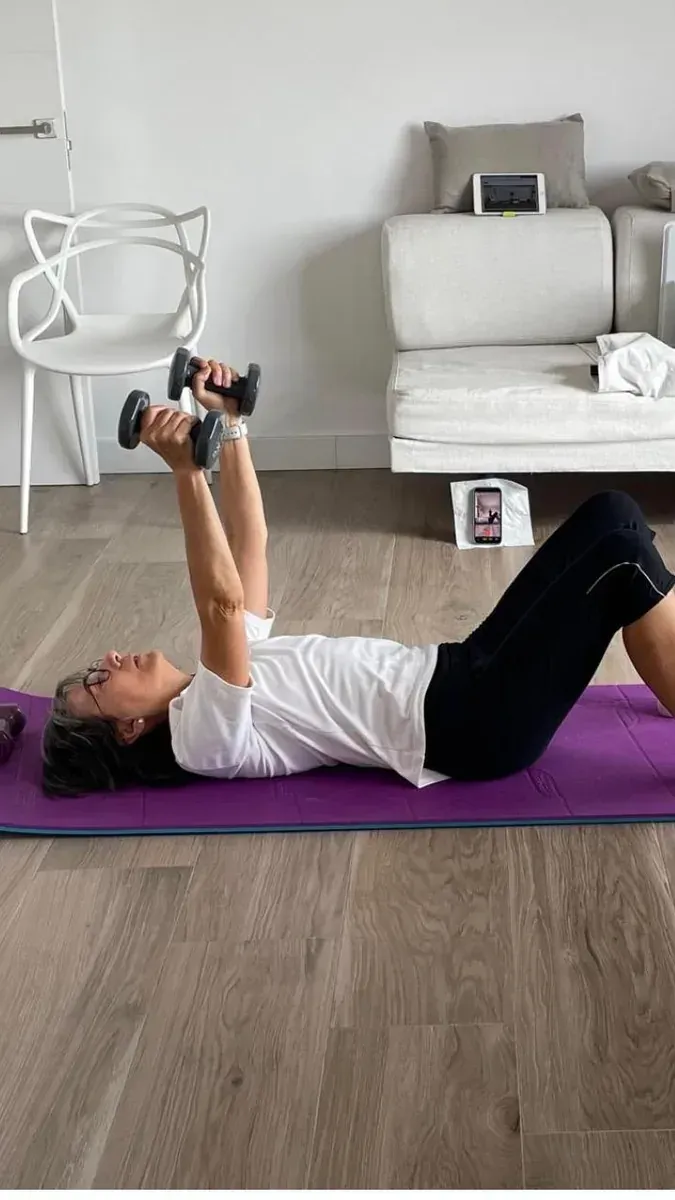 Alumna entrenando fuerza con mancuernas en el salón de su casa, con la clase en la tablet y el móvil grabando