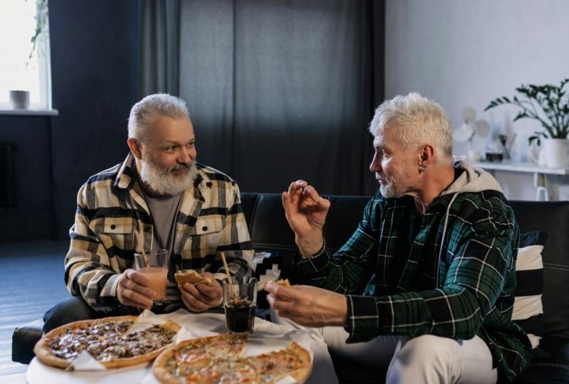 Two elderly male veterans sitting together eating pizza.