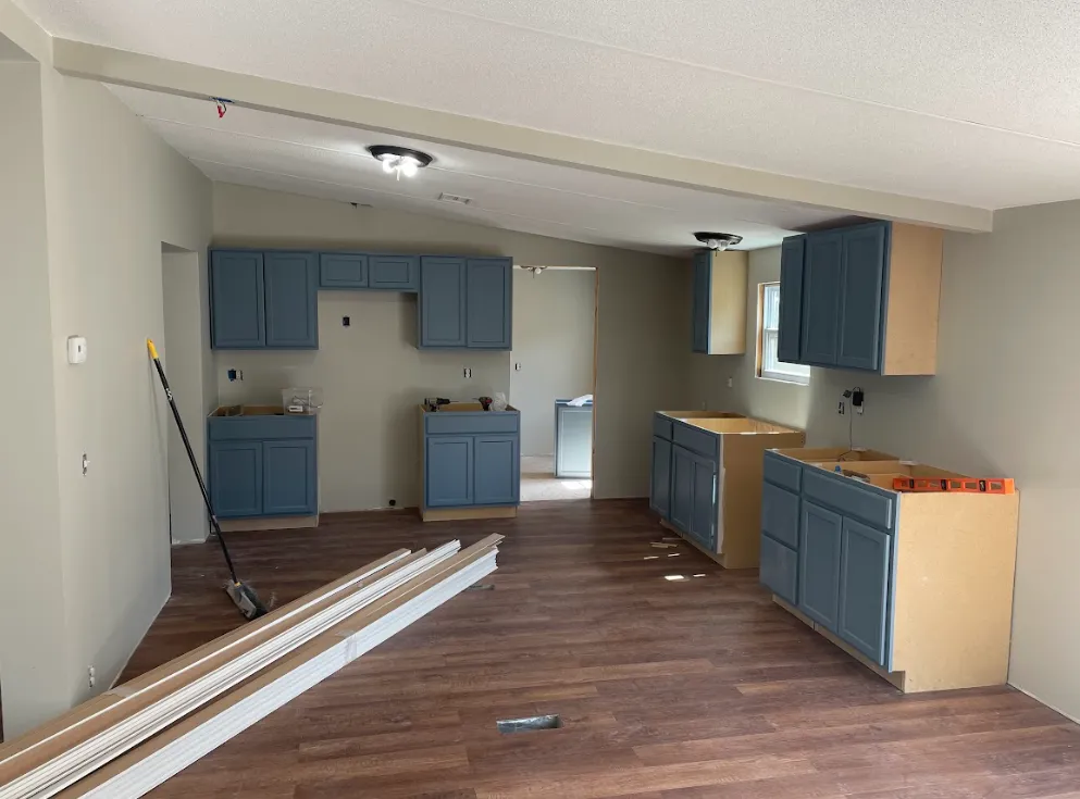 Partially finished kitchen with blue cabinets, brown wood flooring, and light walls. Cabinet doors are missing, and construction materials—including planks and a broom—are scattered on the floor.