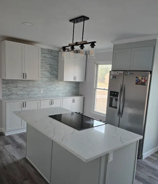 Modern kitchen with white cabinets, gray backsplash, stainless steel refrigerator, large window, and an island featuring a built-in stovetop under a black rectangular light fixture. Light wood flooring.