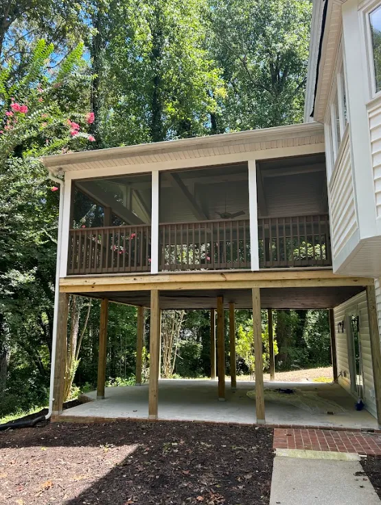 A screened-in porch with a wooden railing is elevated on wooden posts, creating an open area underneath. The structure is attached to a light-colored house surrounded by tall trees and greenery.