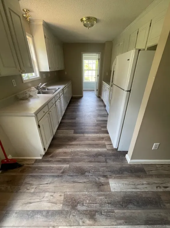 A narrow kitchen with white cabinets, a white refrigerator, a double sink, wood-patterned vinyl flooring, and a windowed door at the far end letting in natural light.