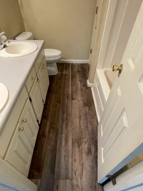 Small bathroom with a double-sink vanity, white cabinets, a toilet, and a shower-tub combination. The floor has a dark wood-like finish, and the walls are a light beige color. The door is partially open.