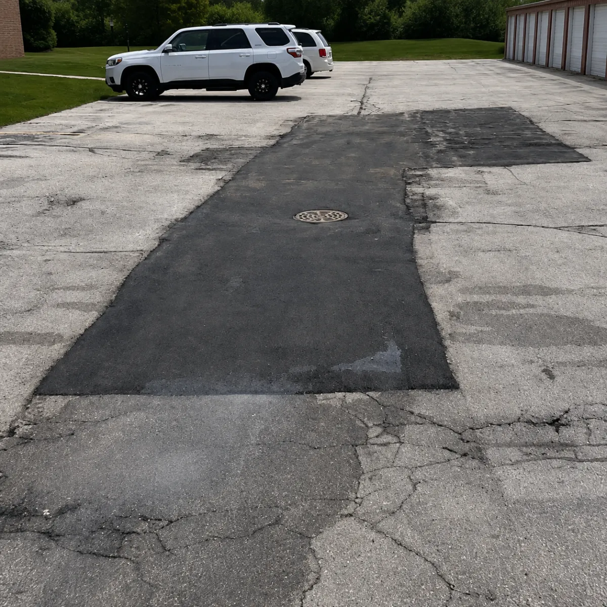 Large asphalt utility cut repair with a manhole cover surrounded by older cracked pavement