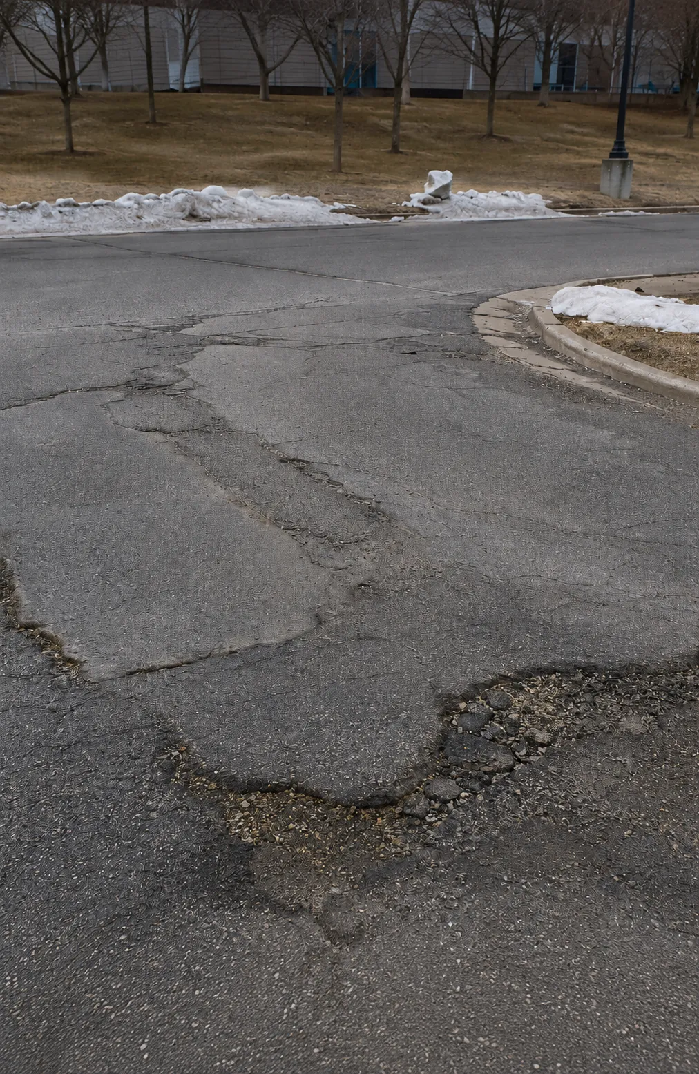 Deteriorated asphalt pavement with cracking, broken edges, and localized failure near a curb during winter