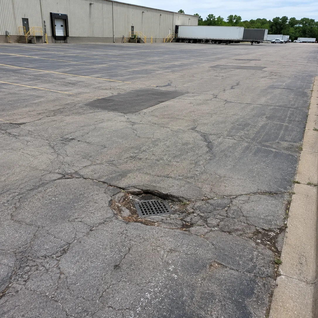 Deteriorated commercial parking lot pavement with cracking and failed asphalt around a storm drain.