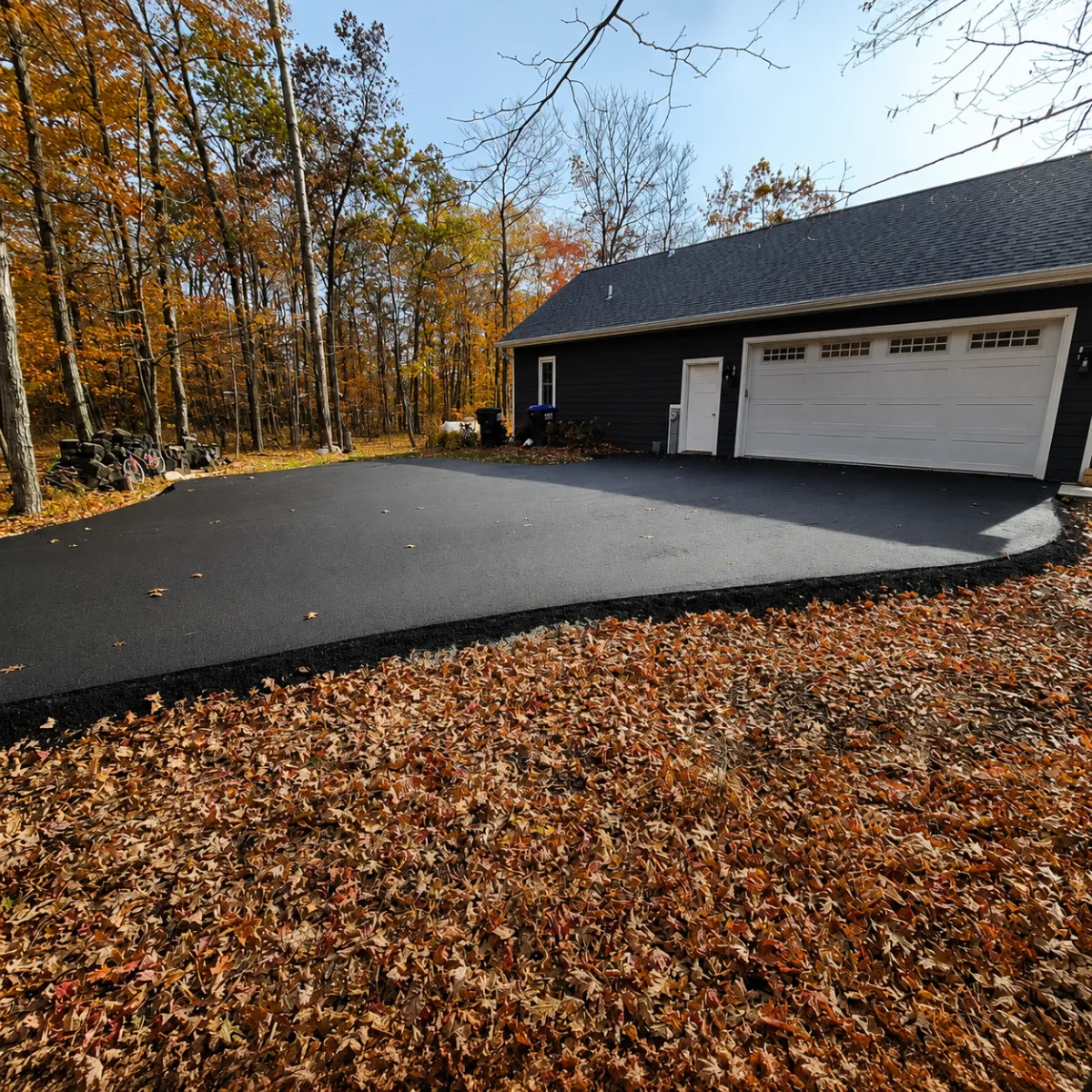 Newly paved residential asphalt driveway leading to a detached garage surrounded by fall trees.