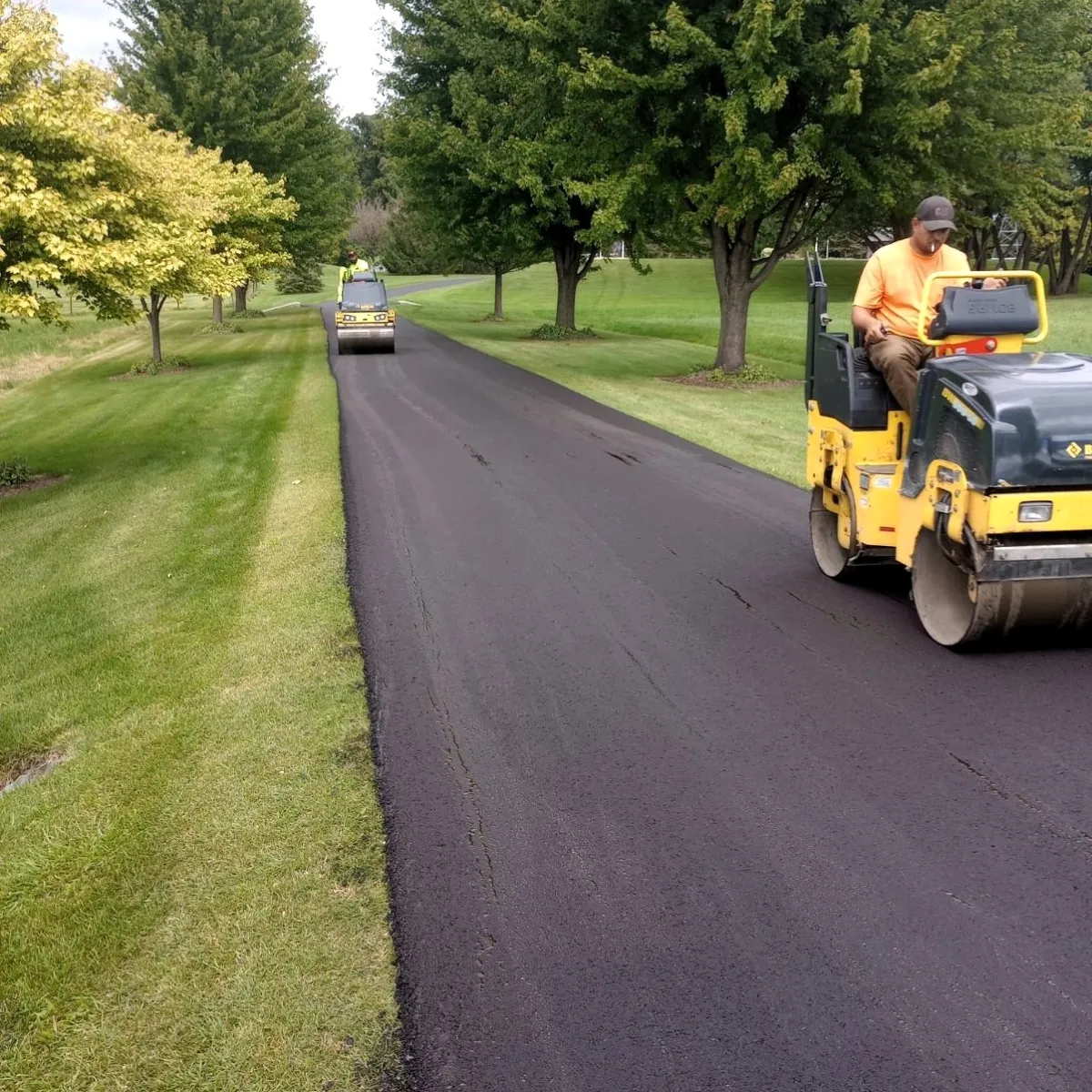 Asphalt rollers compacting a freshly paved residential driveway bordered by grass and trees.